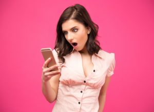 Surprised woman using smartphone over pink background. Wearing in shirt