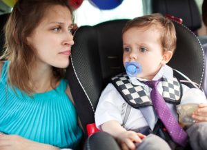 Young mother looking at the dear little son with pacifier and bottle sitting in child safety seat in the car