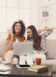 Two excited women shopping online with credit card and laptop sitting on sofa at home, copy space