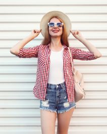 Happy smiling young woman posing in summer round straw hat, checkered shirt, shorts on white wall background