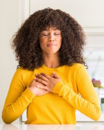 African american woman wearing yellow sweater at kitchen smiling with hands on chest with closed eyes and grateful gesture on face. Health concept.