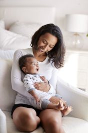 Young adult mother sitting in an armchair in her bedroom, holding her three month old baby son in her arms and looking down at him smiling, vertical