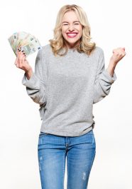 Super excited young woman in grey sweater and jeans holding bunch of Euro banknotes, clinching fists, celebrating winning lottery. Ecstatic woman holding lots of money, isolated on white background.