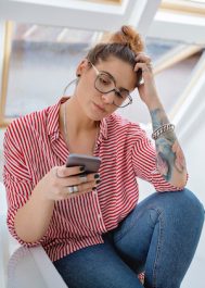 Lovely woman with eyeglasses in red and white shirt, sitting in the kitchen during breakfast using her phone.