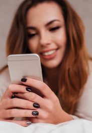 Smiling brunette woman using mobile phone while lying in bed at home, focus on phone